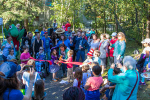 An adult and child use big scissors to cut a long red ribbon while a crowd surrounds them in the park.