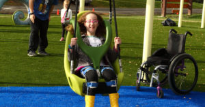 A girl smiles while swinging. A wheelchair sits to the side of the swing.